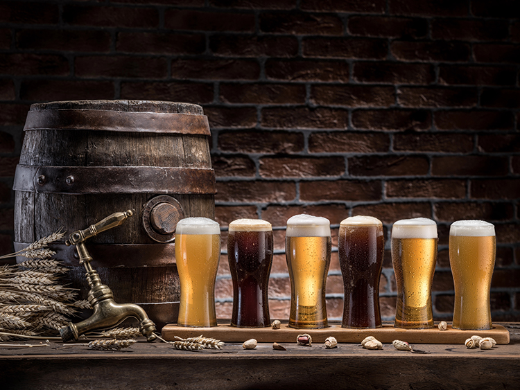 Craft beer tasting flight with six beers served on a wooden paddle beside a brewery barrel.